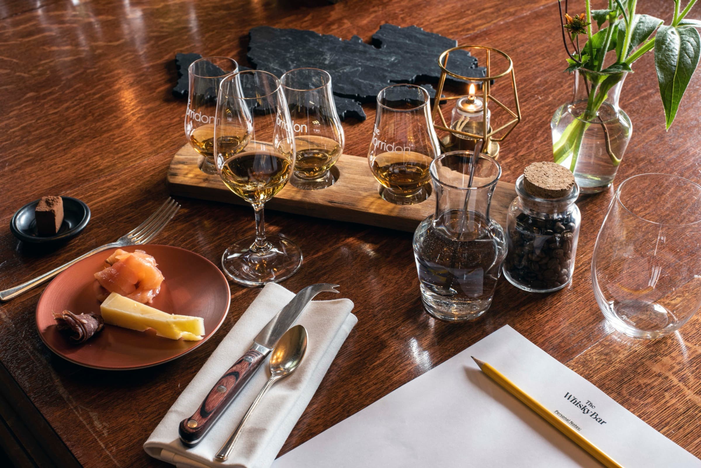 A whisky tasting flight on a dark wood table — three filled Glencairn glasses, a candle in a brass lantern, a plate of smoked salmon and cheese.
