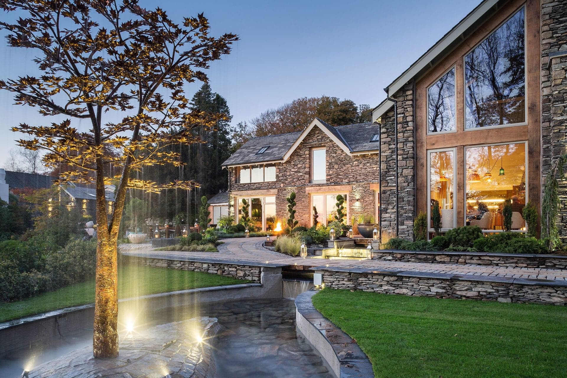 The Gilpin's main house at twilight, lit windows and a stone-and-timber façade behind a small water feature in the foreground.