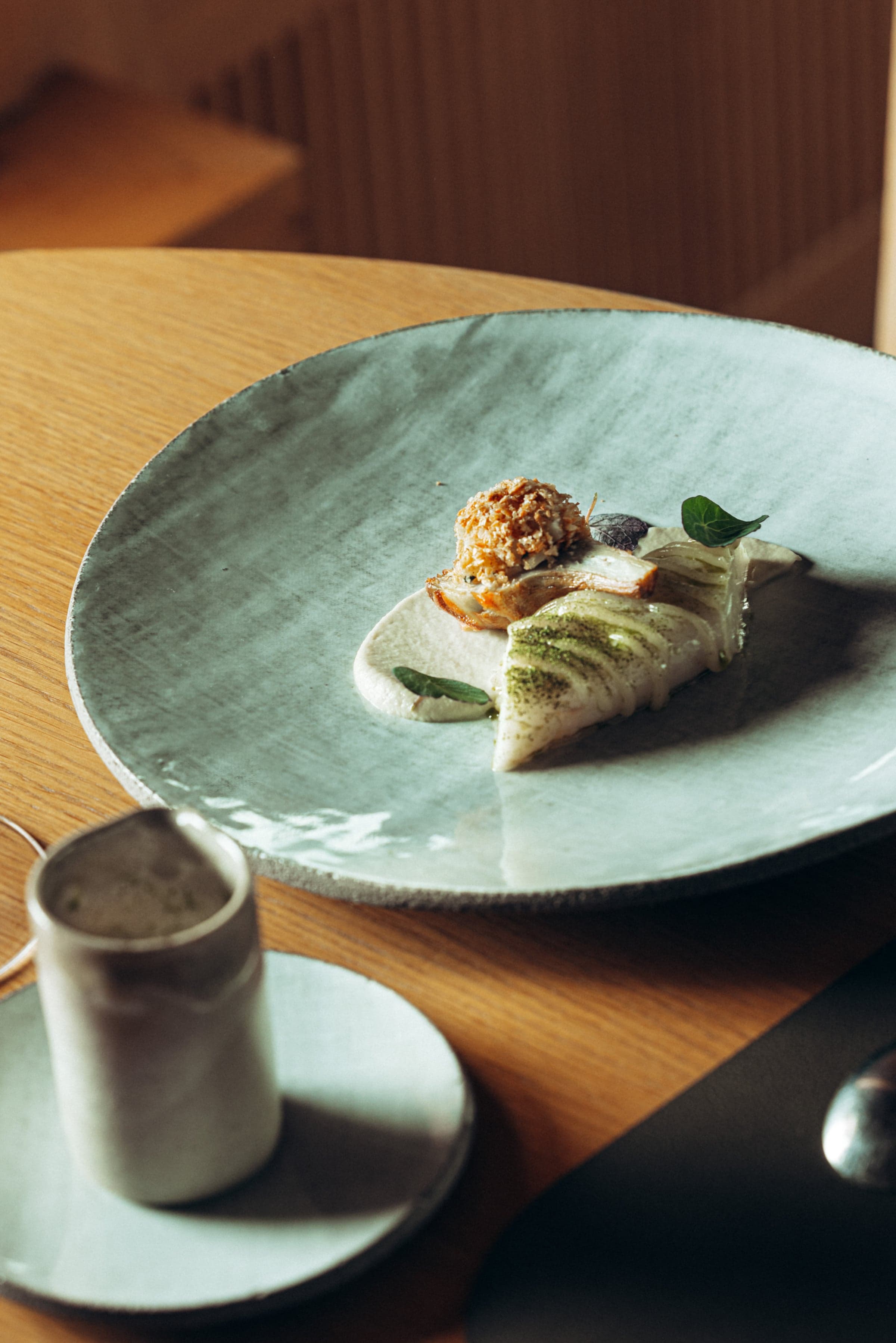 A pale-glazed plate set in side-light, a fillet of fish with green leaves and a small mound of toasted grain.