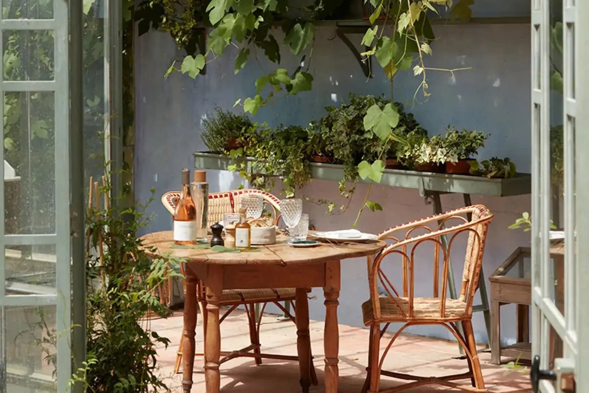 A small round table set inside an orangerie, a bottle of rosé and crystal glasses among the vines, light filtering through the open doors.