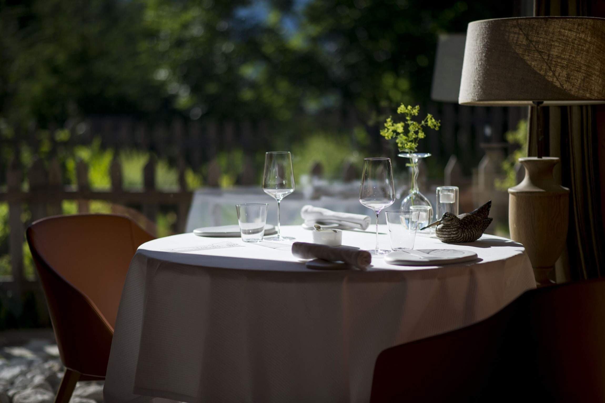 A round table for two laid in white linen, two stemmed glasses and a simple bud vase, set against the open window of the chalet dining room with summer-green pasture and a wooden fence beyond.