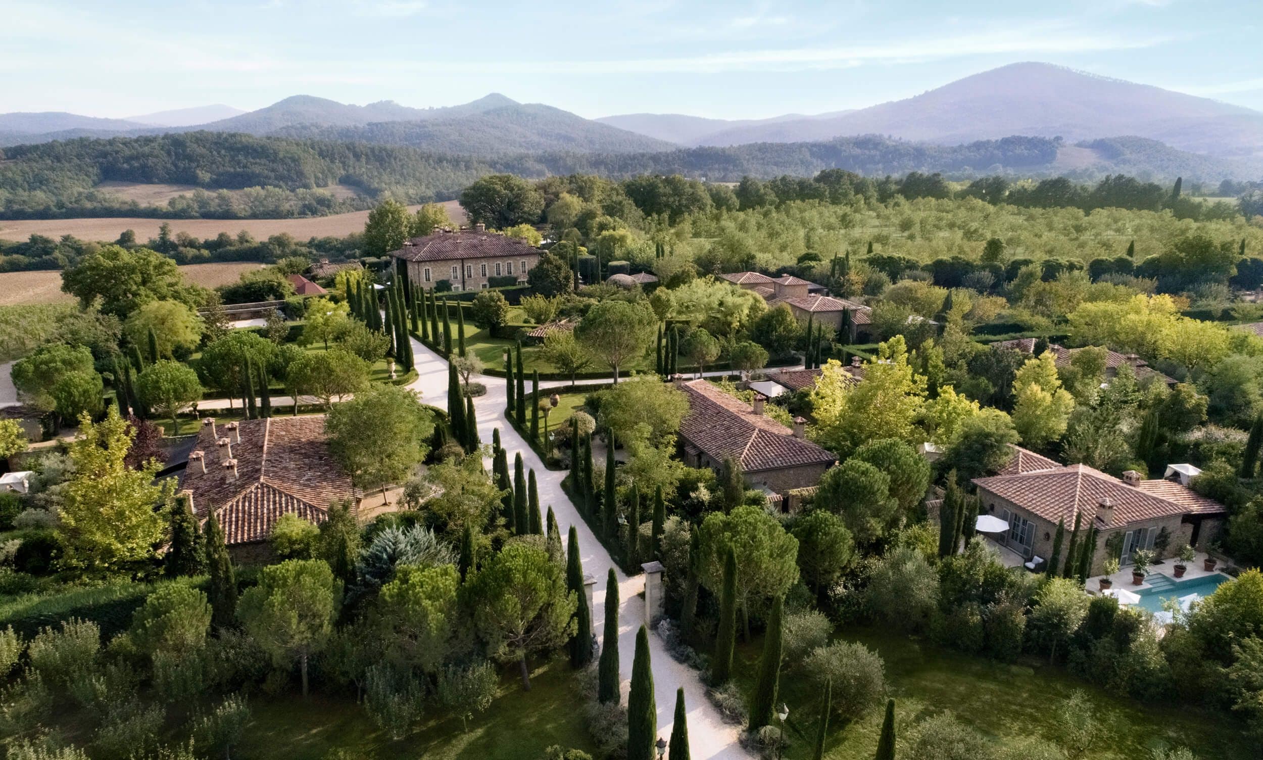 An aerial view of Borgo Santo Pietro — cypress avenues, terracotta roofs and a thirteenth-century villa, the Tuscan hills rising beyond.