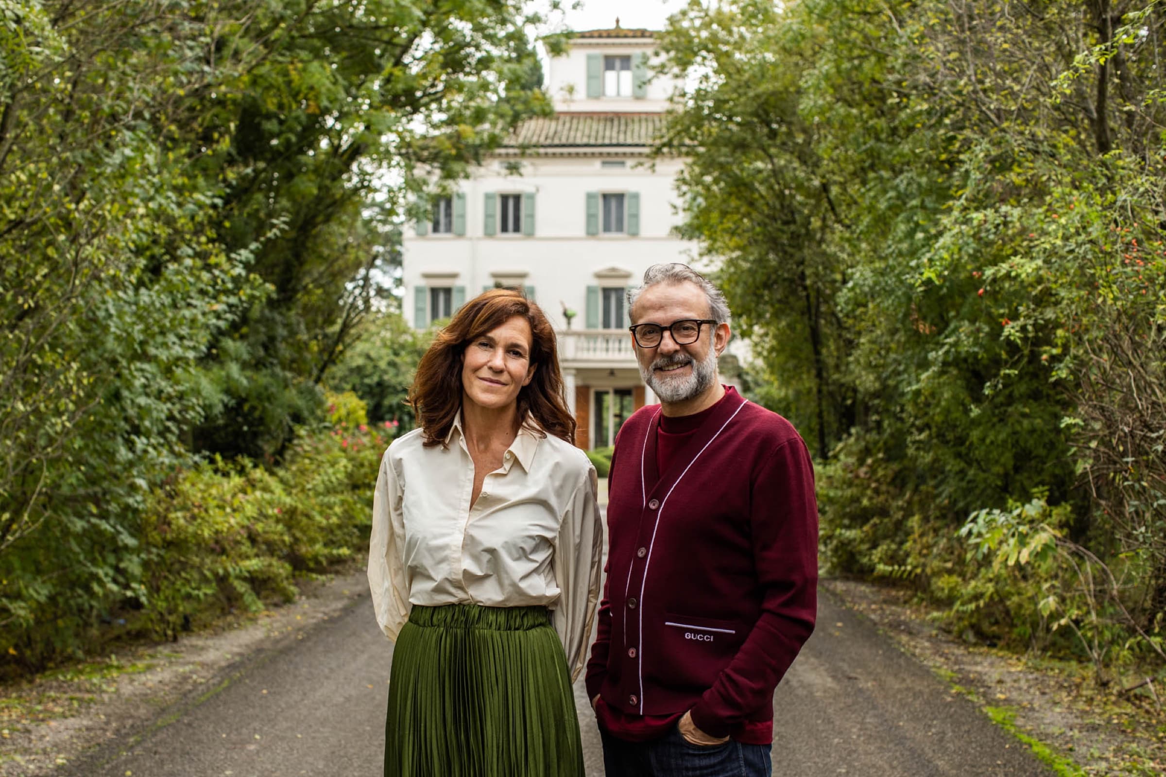 Massimo Bottura and Lara Gilmore standing on the gravel drive in front of the cream-painted villa, hedges and trees framing the house behind them.