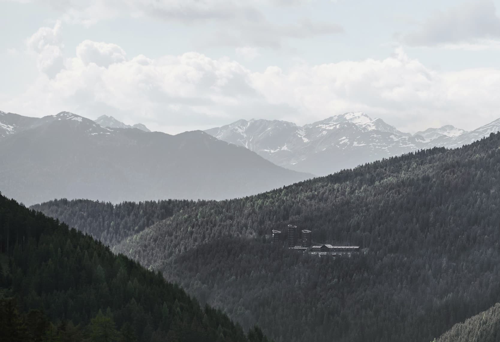 Forestis seen from across the valley — the dark roofs of the historic timber villa held inside dense forest, with a long line of snowed Dolomite peaks behind, under a still afternoon sky.