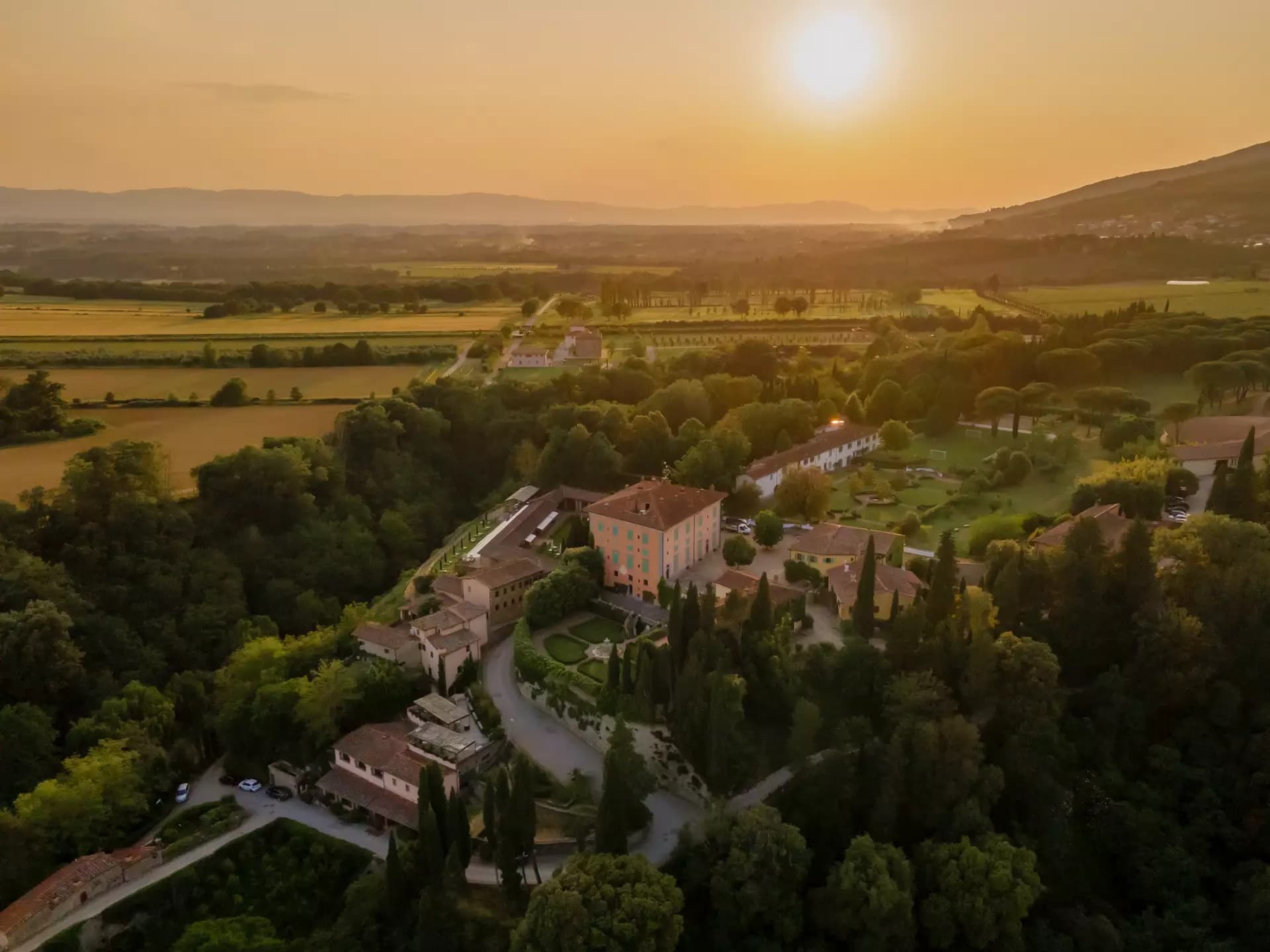 An aerial view of Il Borro at sunset — the rose-coloured villa and its formal Italian garden on a wooded ridge above the Valdarno valley.
