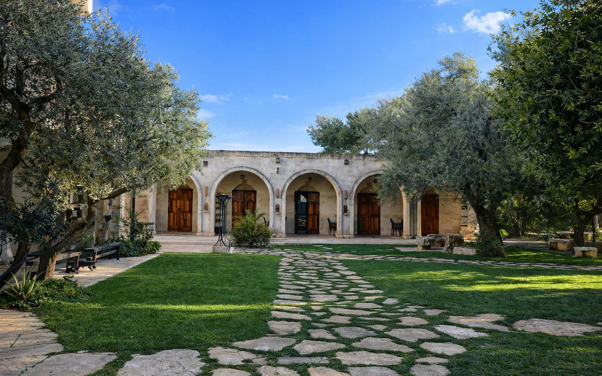The arched stone façade of Masseria Le Lamie at midday, a flagstone path crossing the courtyard between two ancient olive trees.