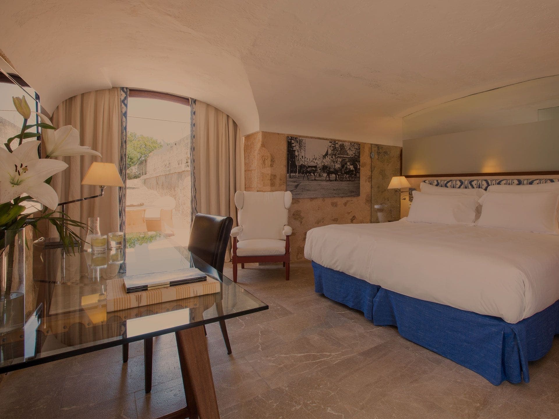 A Doble Fortaleza bedroom under a vaulted stone ceiling — white linen, a sepia photograph mounted on the rough back wall, a tall arched window onto the terrace.