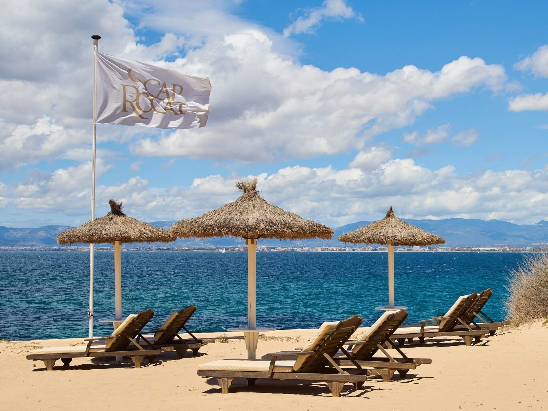 The hotel's small private beach at the Sea Club — straw parasols, wooden loungers, a Cap Rocat flag in a light breeze.