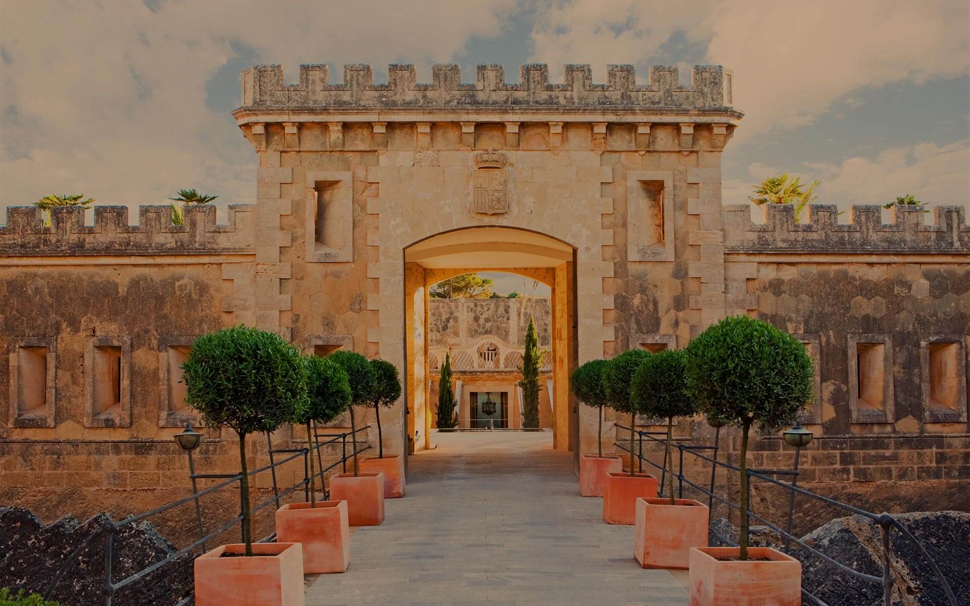 The crenellated stone gate of Cap Rocat at sunset, an arched opening flanked by olive topiaries in terracotta planters.