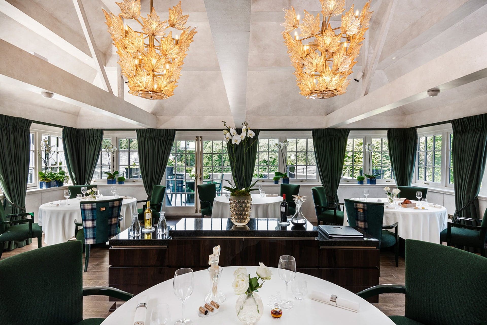 The dining room at The Glenturret Lalique — round tables in pressed white linen on dark herringbone parquet, dark green velvet curtains, dark wood servery, and two large gold-lustre Champs-Élysées Lalique chandeliers hung from the vaulted plaster ceiling.