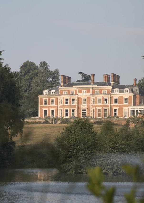 The Georgian house seen from the pleasure grounds at midday, lawns and tall trees in the foreground.