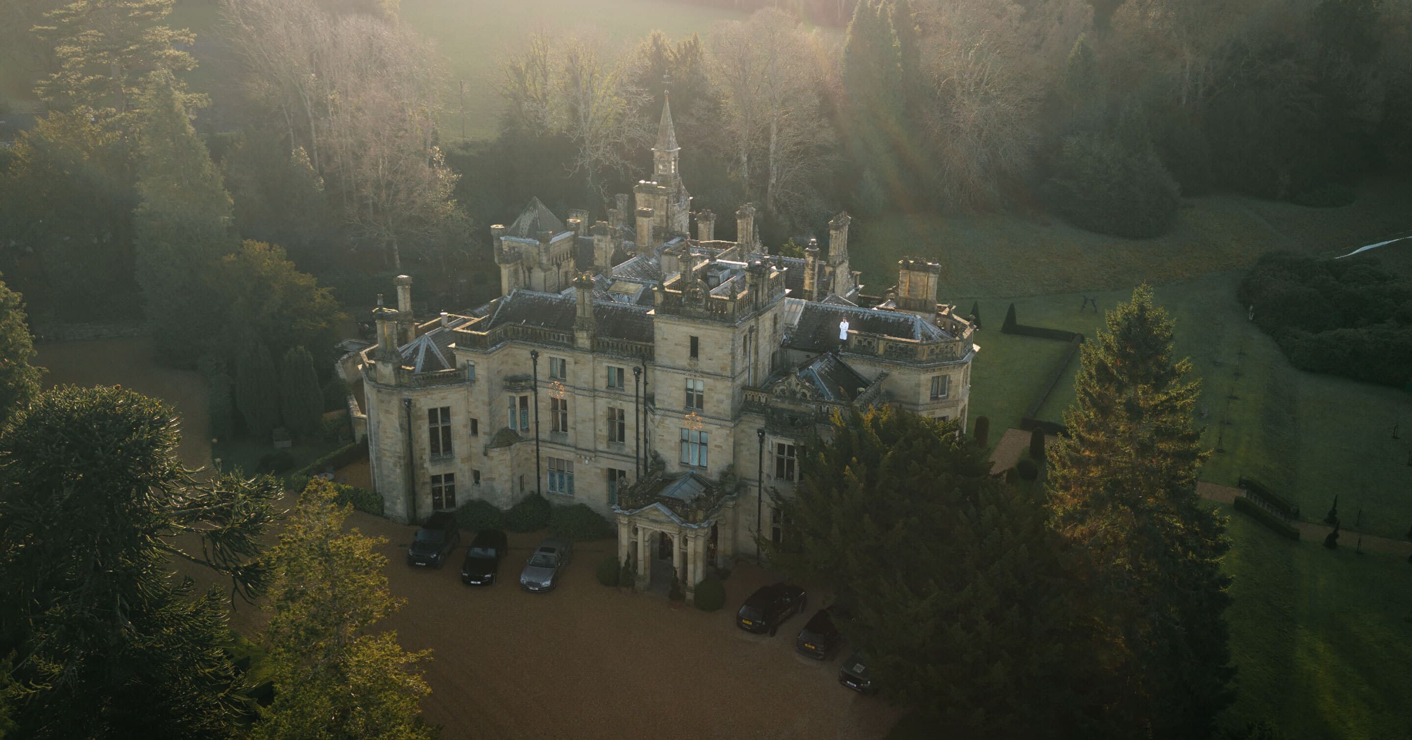 An aerial of the Hall at first light, the Victorian roofline in soft gold against the surrounding woodland.