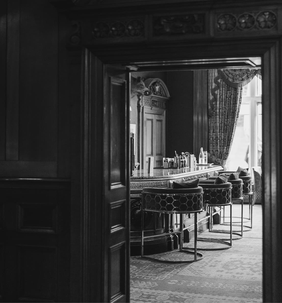 The whisky lounge at Palé Hall in black and white, seen through a wood-panelled doorway, low stools at the marble bar.