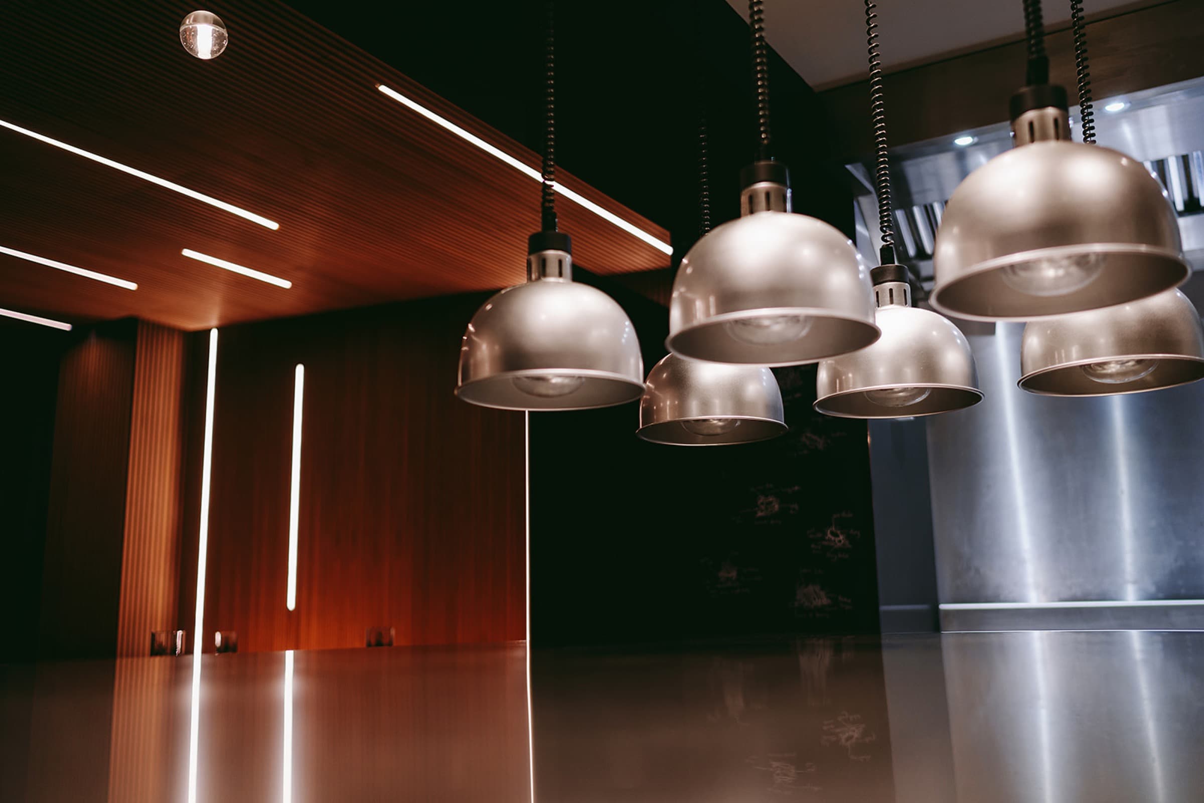 The dining-room counter at Home — a row of brushed-steel pendant lamps lowered over a polished metal pass, a wall of warm timber slats lit with thin vertical strips of light, the open kitchen beyond.