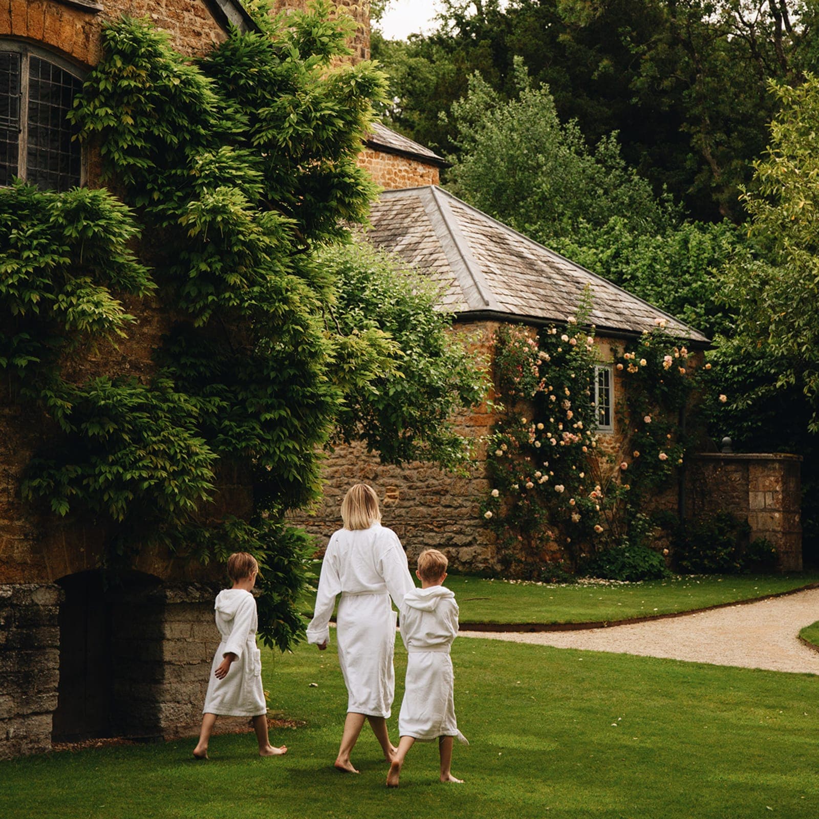 A family in white robes walking through the gardens at Hadspen, past a stone tower covered in climbing roses.
