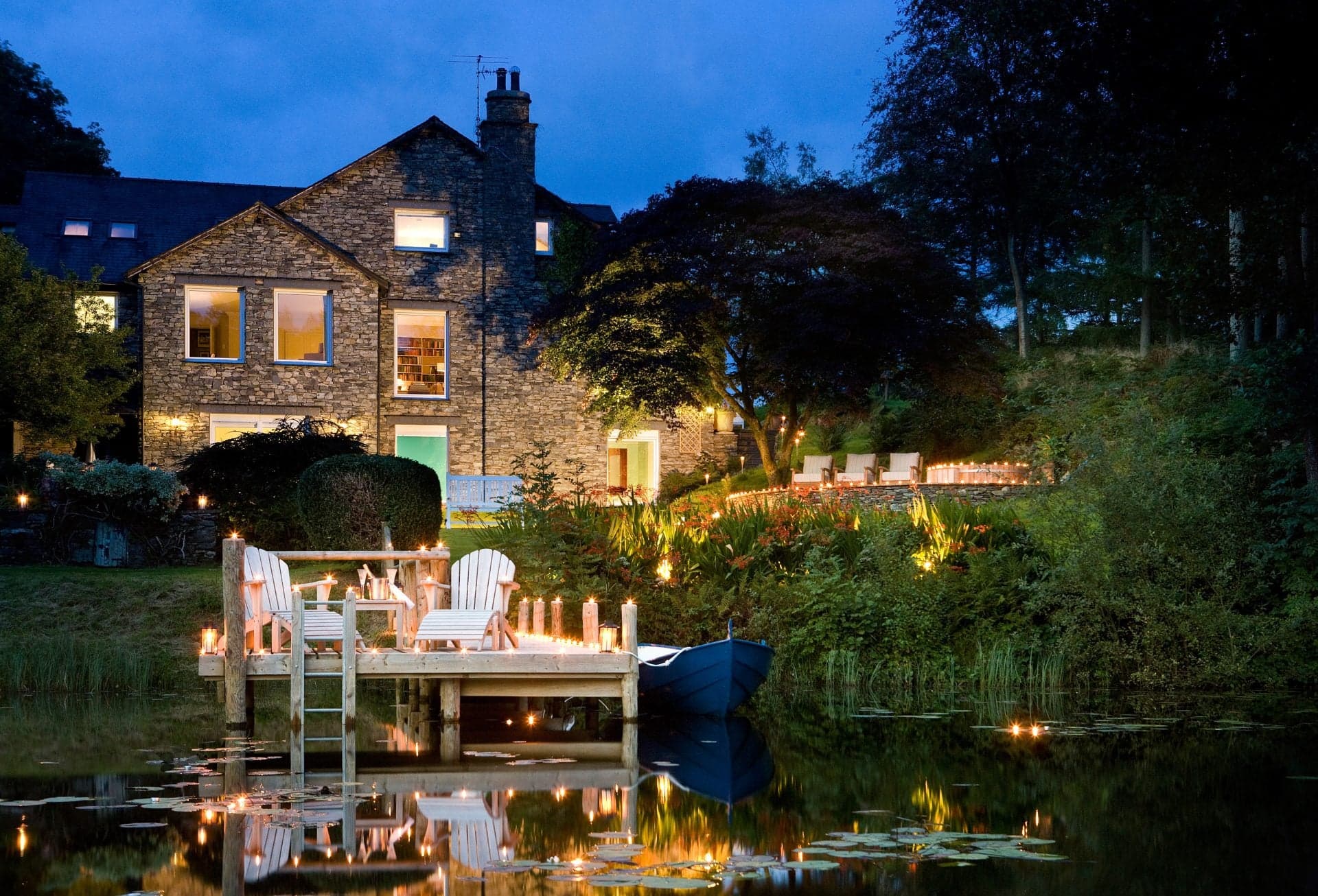 The Lake House at night, two Adirondack chairs and lit candles on a small wooden dock with the still tarn reflecting the lamplit windows above.