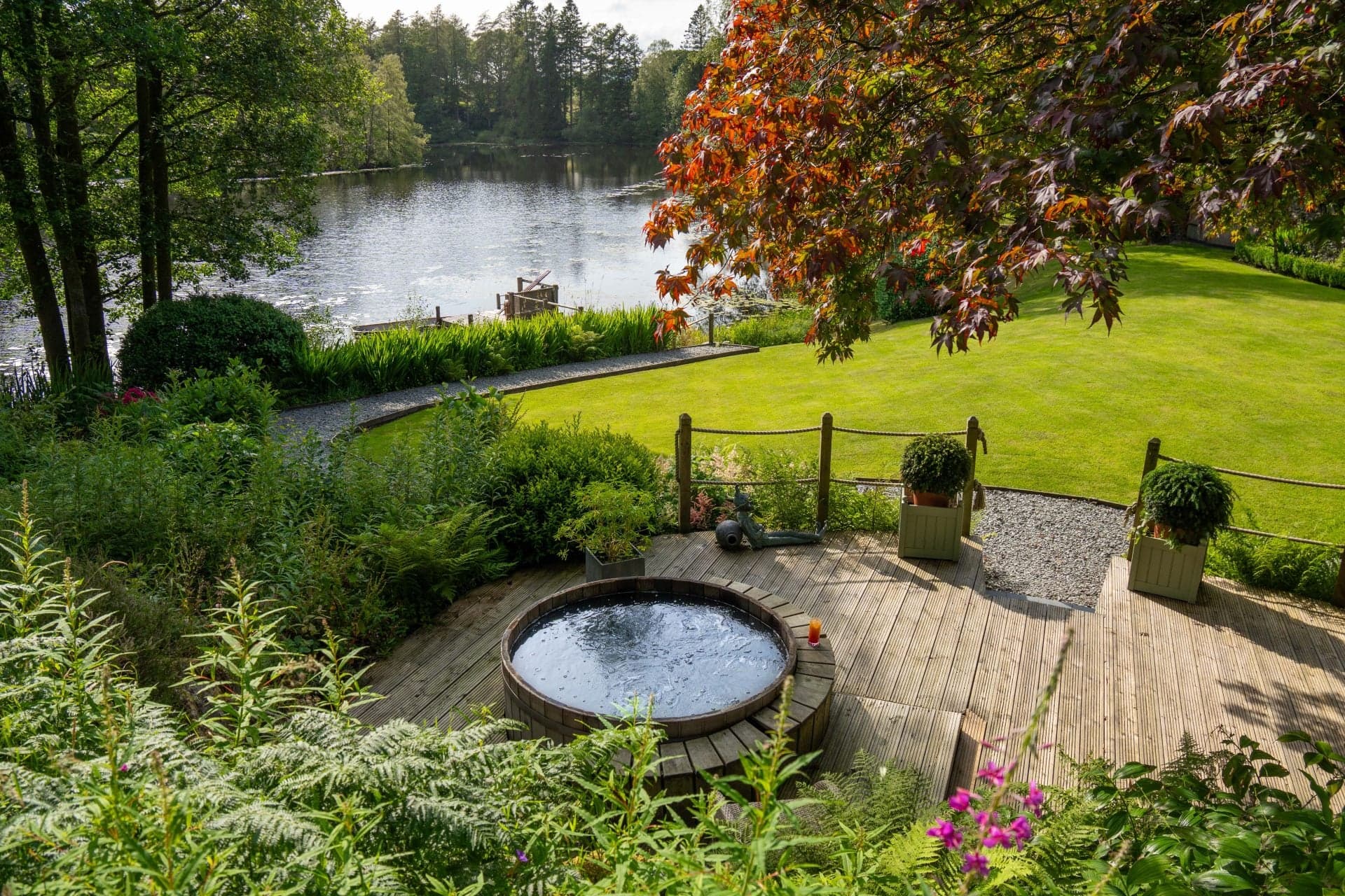A wooden hot tub set on a deck above a tarn, autumn foliage and a small jetty in the background.