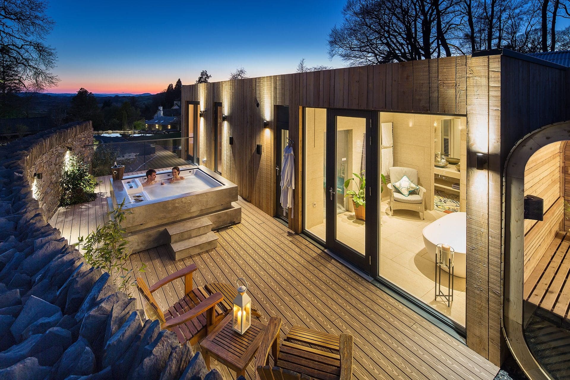 A private spa lodge at twilight, a cedar hot tub on a wooden deck with floor-to-ceiling glass behind, lit from within.