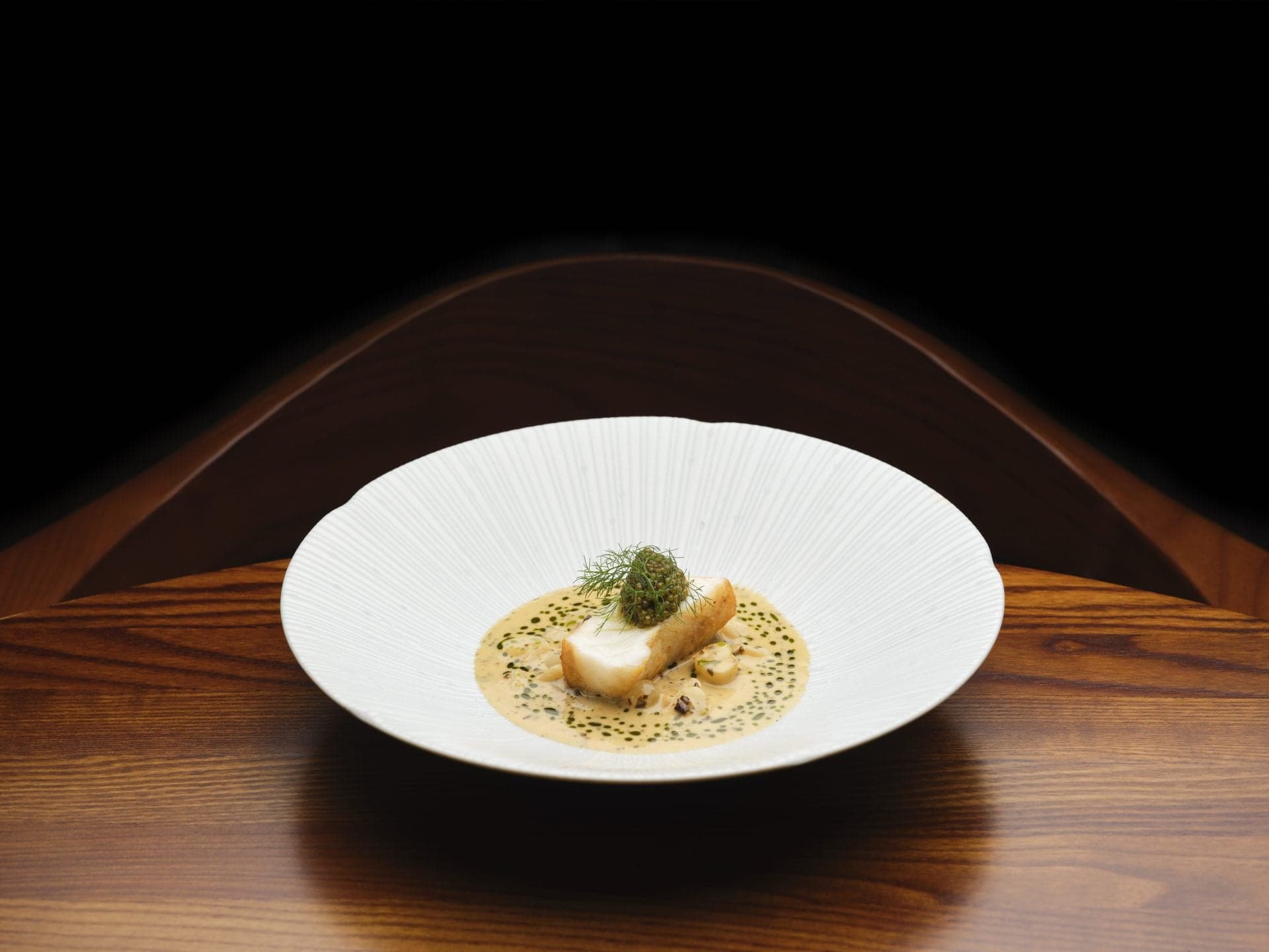 A pale fluted bowl on a dark wooden table, a fillet of monkfish set in a green-flecked cream sauce with a small mound of caviar above.